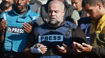L'immagine mostra un gruppo di persone in piedi che sembrano partecipare a un momento di preghiera o riflessione. In primo piano, un uomo indossa un giubbotto con la scritta "PRESS" ed ha una fasciatura su un braccio. Gli altri membri del gruppo, che indossano abbigliamento simile, appare coinvolti nell'atto di pregare, con le mani sollevate. L'atmosfera sembra essere solenne e carica di emotività.