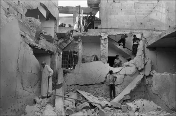 L'image montre un bâtiment en ruines, visiblement endommagé par un conflit ou un désastre. Les murs sont effondrés et des débris jonchent le sol. Plusieurs personnes se trouvent dans l'espace, certains semblent fouiller parmi les débris, tandis que d'autres observent la scène depuis un étage supérieur. L'ambiance est sombre et témoigne des conséquences dévastatrices sur la vie humaine et les infrastructures.