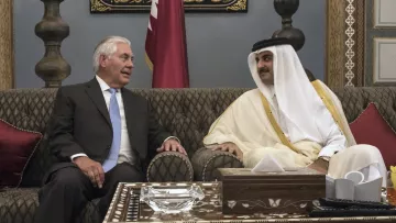 L'image montre deux hommes assis dans un cadre élégant. L'un d'eux porte un costume noir avec une cravate claire, tandis que l'autre est habillé d'une tunique traditionnelle blanche et d'un manteau beige. Ils semblent engagés dans une conversation conviviale, entourés de décorations raffinées, y compris des coussins rouges et des ornements sur la table. L'atmosphère est de camaraderie et de diplomatie.