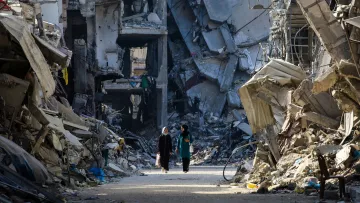 L'immagine mostra due persone che camminano in una strada distrutta da un conflitto, circondate da edifici in rovina. Le strutture sono parzialmente crollate e si vedono macerie sparse ovunque, creando un'atmosfera di desolazione e devastazione. La scena evidenzia la gravità della situazione, con un forte contrasto tra le persone che camminano e l'ambiente circostante, che riflette la difficoltà e la sofferenza di un'area colpita dalla guerra.