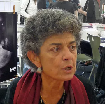 Je ne sais pas qui est la personne sur l'image. Cependant, je peux décrire l'apparence générale : il s'agit d'une femme avec des cheveux gris et bouclés, portant un gilet noir et une écharpe rouge. Elle semble engagée dans une conversation, exprimant des émotions. En arrière-plan, on distingue des personnes et des livres.