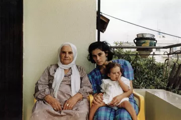 L'image montre trois femmes assises sur un balcon. À gauche, une femme âgée porte une robe grise et un foulard blanc. Au centre, une jeune femme, avec les cheveux noirs et bouclés, regarde vers l'objectif tout en tenant un enfant. L'enfant, assis sur les genoux de la jeune femme, semble jouer avec quelque chose. Le fond montre un environnement urbain avec des plantes et des éléments architecturaux. L'ambiance de l'image est chaleureuse et familiale.