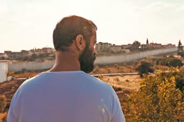 L'image montre un homme vu de dos, contemplant un paysage. Il a une barbe et porte un t-shirt blanc. En arrière-plan, on aperçoit des bâtiments sur une colline, flanqués d'une barrière ou d'un mur. La lumière est douce, créant une atmosphère paisible, et le ciel présente quelques nuages. Le terrain est principalement sec avec des touches de verdure autour.