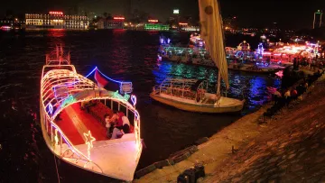 L'immagine mostra una scena notturna su un fiume, con diverse barche decorate con luci colorate. Le imbarcazioni si trovano in una zona animata, e il riflesso delle luci sull'acqua crea un'atmosfera vivace e festosa. Sullo sfondo, si possono vedere edifici illuminati, che contribuiscono ulteriormente all'ambiente festivo della scena. Alcune persone sono sedute lungo la riva, godendosi la vista e l'atmosfera.