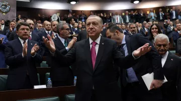 الصورة تظهر رئيس تركيا، رجب طيب أردوغان، standing في مجلس يتكون من عدد كبير من الشخصيات. يبدو أنه يتحدث أو يقدم خطابًا، حيث يرفع ذراعيه بشكل يعبر عن التواصل أو الاستجابة. خلفه، يوجد مجموعة من الحضور الذين يصفقون أو يستمعون له، مما يوحي بأجواء من التفاعل والحماس. ترتدي الشخصيات خلفه ملابس رسمية، ويبدو أنهم في جلسة برلمانية أو حدث سياسي.