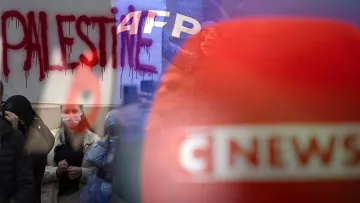 L'image présente une scène de manifestation. On peut voir un graffiti rouge avec le mot "PALESTINE" écrit en grandes lettres. En arrière-plan, on distingue des personnes rassemblées, certaines portant des masques. Des emblèmes de médias, comme "AFP" et "CNEWS", sont également visibles, suggérant une couverture médiatique de l'événement. L'atmosphère semble être chargée d'émotion et de revendication.