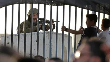 L'image montre un soldat armé stationné derrière une barrière ou un grillage. Il est en position de vigilance, regardant au loin. En face de lui, un jeune garçon semble gesticuler, tandis que d'autres enfants ou adolescents se trouvent à proximité. L'environnement semble tendu, reflétant une situation de contrôle ou de conflit. La lumière du soleil indique un moment de la journée où l'activité est encore présente.