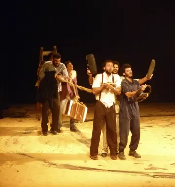 L'image montre un groupe de personnes sur une scène de théâtre, en train de créer une sorte de performance. Ils semblent emporter différents objets, comme des chaises et une valise, en formant une sorte de procession. La scène est éclairée de manière à mettre en évidence leur mouvement, tandis que le fond est sombre, ce qui attire l'attention sur les acteurs. L'ensemble donne une impression dynamique et dramatique, évoquant une histoire ou une interaction entre les personnages.