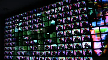 L'immagine mostra un ampio muro composto da molteplici schermi televisivi, ognuno dei quali visualizza immagini colorate e astratte. Le immagini si sovrappongono e si mescolano creando un effetto visivo vivace e dinamico. La combinazione di colori e forme sembra generare un'atmosfera futuristica e artistica, dando l'impressione di un'opera d'arte digitale contemporanea.