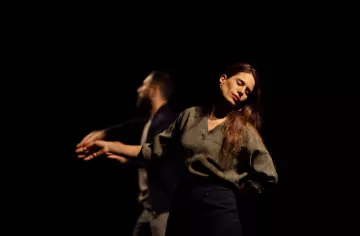 Deux danseurs se déplacent sur scène dans une lumière tamisée. Une femme, les yeux fermés et une expression de sérénité, se tient légèrement en avant, tandis qu'un homme, derrière elle, semble effectuer un mouvement fluide avec ses bras. L'atmosphère est intimiste et expressive.