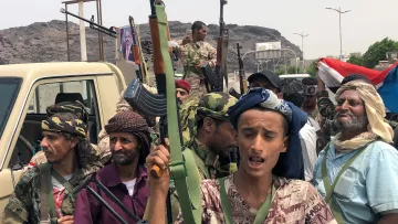 L'image représente un groupe de personnes armées, apparemment réunies dans un contexte de lutte ou de manifestation. Ils portent des vêtements typiques et semblent déterminés, avec des regards engagés. En arrière-plan, on peut apercevoir des montagnes et une circulation modérée, ainsi qu'un drapeau. Un véhicule est également visible, avec des armes visibles entre les membres du groupe. L'atmosphère suggère une situation de tension ou de conflit.