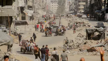 L'immagine mostra una scena di una città con edifici distrutti e rovine visibili. La strada è affollata da persone che camminano e da veicoli, inclusi motocicli e carretti. La polvere sollevata dall'ambiente circostante crea un'atmosfera di desolazione. In lontananza si vedono ulteriori segni di distruzione, con detriti sparsi lungo i lati della strada. L'insieme comunica un forte senso di difficoltà e resilienza in un contesto di crisi.