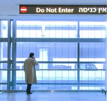 L'image montre un homme debout près d'une grande fenêtre dans un espace intérieur moderne. Il porte un long manteau clair et semble être en train de parler au téléphone. Au-dessus de lui, il y a une signalisation en trois langues, indiquant "Do Not Enter" en anglais et une phrase en hébreu, ainsi qu'un panneau rouge avec un symbole d'interdiction. L'espace est bien éclairé, avec des murs en verre et une ambiance assez minimaliste.
