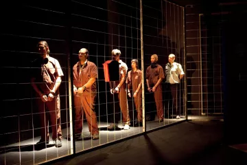 L'image montre un groupe d'hommes disposés en ligne, chacun portant des vêtements de style uniforme. Ils se tiennent derrière un grillage, ce qui crée une impression de séparation ou de détention. L'éclairage est sombre, mettant en valeur les figures des hommes, qui semblent concentrés ou pensifs. L'ambiance générale évoque des thèmes de confinement et d'isolement.