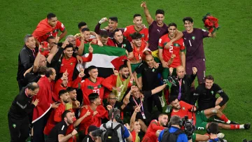 این تصویر نشان‌دهنده خوشحالی و شادی تیم ملی فوتبال مراکش پس از یک پیروزی بزرگ است. بازیکنان و اعضای کادر فنی تیم در حال جشن گرفتن هستند و بعضی از آن‌ها پرچم مراکش را در دستان خود دارند. فضای تصویر پر از انرژی مثبت و شادی است و بازیکنان با لبخند و هیجان در کنار یکدیگر قرار دارند. همچنین تعدادی از عکاسان و اعضای رسانه نیز در حال تصویربرداری از این لحظه مهم هستند.