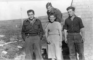 L'image montre quatre personnes se tenant debout à l'extérieur, probablement sur un terrain dégagé. Il y a trois hommes et une femme. Les hommes portent des uniformes militaires ou des vêtements utilitaires, tandis que la femme est habillée simplement dans une robe. L'environnement semble rural, avec une petite bâtisse en arrière-plan et des champs en toile de fond. Tout le monde affiche des expressions neutres ou souriantes, créant une atmosphère de camaraderie.