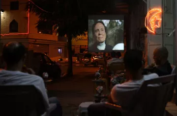 L'image montre une scène nocturne où un groupe de personnes est assis à l'extérieur, observant un film projeté sur un mur. On voit des chaises disposées face à l'écran, et quelques fumées de narguilé flottent dans l'air. L'environnement est animé par des lumières de néon, et on aperçoit des voitures garées et des bâtiments en arrière-plan, créant une ambiance conviviale et détendue.