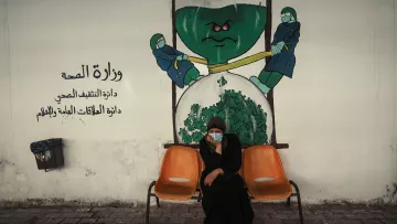 الصورة تظهر امرأة جالسة على كرسيين في منطقة تبدو وكأنها مركز صحي أو مستشفى. تحمل المرأة ملامح الجدية، وتضع كمامة على وجهها. خلفها يوجد جدار م painted يوضح رسماً يجسد فكرة وزارة الصحة، حيث يظهر شخصيتان ترتديان بزة وقاية، تسعيان لشد أو دفع شيء كبير يشبه الخضار أو الملفوف. الألوان المستخدمة في الجدار تعكس موضوع الصحة، مع تركيبات واضحة ورموز مثل القفازات والكمامات.