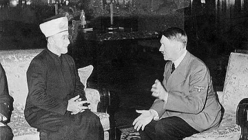 Cette image montre deux hommes assis dans une pièce. L'un d'eux porte un habit traditionnel avec un couvre-chef, tandis que l'autre est vêtu d'un uniforme militaire. Ils semblent être en conversation, gesticulant avec leurs mains. L'environnement est richement décoré, avec des meubles élégants et des éléments de décoration en arrière-plan. L'image évoque un moment de rencontre entre ces deux figures historiques.