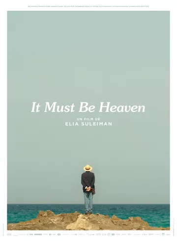 L'image représente une affiche de film intitulée "It Must Be Heaven", réalisé par Elia Suleiman. On y voit un homme de dos, portant un chapeau, se tenant debout sur un rocher au bord de la mer. L'horizon est dégagé, avec un ciel bleu clair. Le titre du film est inscrit en lettres blanches en bas de l'affiche, avec également le nom du réalisateur. L'ensemble de l'image évoque une ambiance contemplative et poétique.