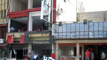 تظهر في الصورة مشهدًا لجزء من مدينة أو منطقة تجارية تتضمن مباني تحتوي على واجهات مدمرة. يوجد بعض الأشخاص الذين يتواجدون في المنطقة، بينما يبدو أن هناك أعمال صيانة أو ترميم جارية. الأرض مبللة ويبدو أن هناك فوضى مرتبطة بأغراض متناثرة. بعض المحلات مغلقة، والفوضى تعكس أجواء معينة من النشاط أو الضرر.