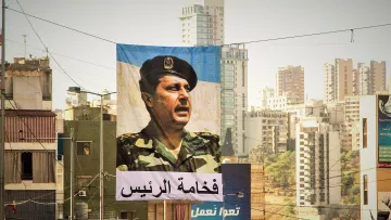 L'image montre une grande bannière suspendue dans une rue urbaine. Sur cette bannière, un portrait d'un homme en uniforme militaire est visible, face à un public. En dessous de l'image, il y a un texte en arabe qui semble faire référence à une autorité ou un leadership. En arrière-plan, on aperçoit des bâtiments modernes et d'autres structures urbaines typiques d'une ville. La lumière est douce, ce qui donne une atmosphère particulière à la scène.