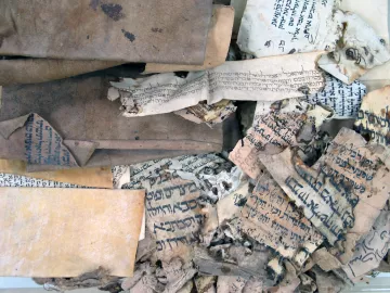 L'image montre un ensemble de vieux manuscrits et de documents enroulés et usés. Les pages sont usées, certaines sont jaunies et déchirées, et on peut voir des inscriptions écrites dans des langues anciennes et probablement des alphabets différents. La texture des papiers varie, certains étant plus épais que d'autres, et la composition générale donne une impression d'antiquité et de fragilité, suggérant leur importance historique.