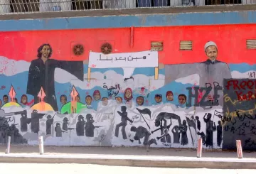 L'image montre une grande fresque murale qui illustre des figures humaines et des symboles de résistance. À gauche, on voit une personne avec de longs cheveux, et à droite, un homme âgé portant un turban. Entre eux, il y a une inscription dans une langue arabe. Le mural est riche en détails, avec des silhouettes de personnes et des flèches, suggérant des thèmes de lutte et d'espoir. Le fond est principalement rouge, créant un contraste fort avec les personnages et les éléments représentés.