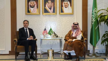 L'image montre deux hommes assis côte à côte dans un cadre formel. À l'arrière-plan, il y a trois portraits encadrés de dirigeants saoudiens. Sur une table entre eux, se trouvent des éléments décoratifs. L'un des hommes porte un costume sombre et l'autre est habillé en tenue traditionnelle saoudienne. À droite, on aperçoit un drapeau saoudien. L'atmosphère semble officielle et diplomatique.