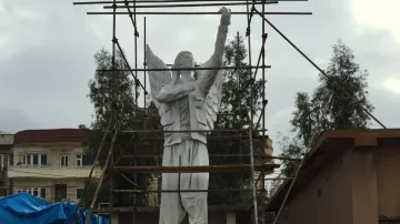 L'image montre une statue de grande taille, représentant un personnage avec des ailes, qui semble être en train d'être rénovée ou construite. Des échafaudages entourent la statue, indiquant qu'elle est encore en travaux. En arrière-plan, on peut entrevoir des bâtiments et une structure en toile bleue, suggérant une ambiance urbaine. Le ciel est nuageux, ce qui donne une atmosphère un peu mélancolique à la scène.