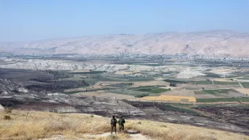 در این تصویر، دو نفر در حال ایستادن بر روی یک تپه یا ارتفاع هستند و به منظرۀ وسیع و زیبای زیر پای خود نگاه می‌کنند. در زیر، دشت‌ها و زمین‌های کشاورزی با رنگ‌های مختلف دیده می‌شوند. دامنه‌های کوه‌ها در دوردست قرار دارند و آسمان آبی و صاف به چشم می‌خورد. فضا به نظر آرام و دلنشین می‌رسد و نشان‌دهندهٔ مناظر طبیعی و انسانی در منطقه‌ای کوهستانی است.