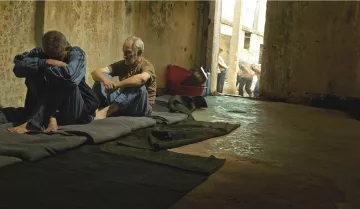 L'image montre un espace intérieur sombre et désolé, probablement une cellule ou un lieu de détention. À l'avant, deux hommes sont assis sur des couvertures posées sur le sol. L'un semble pensif et acculé, tandis que l'autre est plus détendu et semble essayer de communiquer ou de réconforter. En arrière-plan, d'autres personnes sont visibles, marchant en file. L'éclairage est faible, ce qui contribue à une atmosphère de tristesse et de confinement. Les murs semblent usés et abandonnés, renforçant l'impression de désespoir.