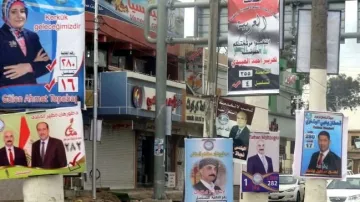 L'image montre une rue animée bordée de panneaux électoraux. On peut y voir plusieurs affiches de campagne, chacune présentant différents candidats avec leurs noms et numéros associés, ainsi que des logos de partis politiques. Les couleurs des affiches varient, attirant l'attention des passants. L'environnement urbain semble typique d'une zone commerçante, avec des boutiques et des enseignes visibles en arrière-plan.