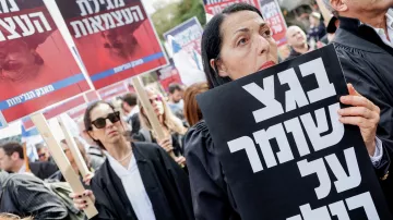 Nell'immagine si vede una manifestazione di persone vestite con abiti neri, probabilmente avvocati, che tengono cartelli. In primo piano, una donna tiene un grande cartello con un messaggio ben visibile. Sullo sfondo, ci sono altre persone con cartelli, in un'atmosfera di protesta. Le persone sembrano serie e impegnate nella loro causa. L'ambiente è quello di una manifestazione pubblica, con molta partecipazione.