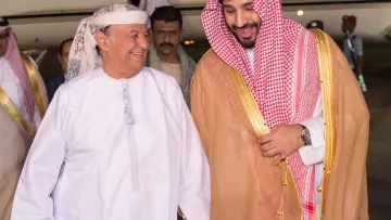 في الصورة، يمكن رؤية رجلين يتحدثان معاً بمرح. يرتدي أحدهما زياء تقليدية عربية مكونة من جبة بيضاء وعقال، بينما يرتدي الآخر الملابس السعودية التقليدية، تشمل العباءة والكوفية. يظهر خلفهم طائرة خاصة، مما يشير إلى وصولهم إلى مكان محدد. تبدو الأجواء مفعمة بالترحاب والود.