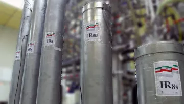 تظهر في الصورة مجموعة من الأسطوانات المعدنية، وكل منها يحمل ملصق مكتوب عليه "IR8" أو "IR8s" مع رمز علم دولة إيران. خلف هذه الأسطوانات، يمكن رؤية مجموعة من الأنابيب والتجهيزات الأخرى، مما يشير إلى أنها قد تكون جزءًا من منشأة صناعية أو مركز بحث علمي. الجو العام في الصورة يعكس بيئة تقنية عالية ومجهزة بأجهزة متطورة.