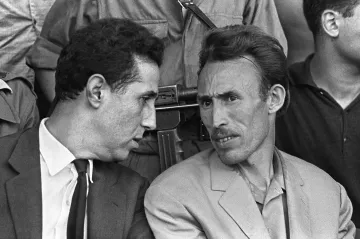 Cette image en noir et blanc montre deux hommes en discussion. L'un porte un costume sombre avec une cravate, tandis que l'autre est habillé de manière plus décontractée avec une veste claire. Ils semblent en train d'échanger des informations ou des réflexions, affichant des expressions attentives. En arrière-plan, on peut apercevoir d'autres personnes, et un fusil est visible, suggérant un contexte de tension ou de sécurité. L'image capte une moment d'intimité entre les deux hommes.