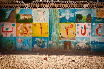 L'image montre un mur décoré de peintures colorées. Les motifs incluent des formes géométriques, des fleurs et des éléments abstraits. Les couleurs vives comme le bleu, le vert, le jaune et le rouge créent un contraste attrayant. Le bas du mur semble légèrement abîmé par le temps, et le sol est recouvert de petits débris, ce qui donne un aspect un peu désordonné à l'ensemble. Cette scène évoque un environnement créatif, peut-être dans un espace communautaire ou scolaire.