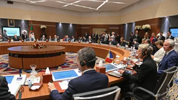 تظهر الصورة اجتماعًا يُعقَد في قاعة مؤتمرات، حيث يجلس العديد من المشاركين حول طاولة دائرية. تتواجد شاشات عرض أمام بعض الأشخاص، وتبدو القاعة مزينة ببعض الزهور. يُلاحظ وجود عدد من الحضور في الخلفية، مما يشير إلى أهمية الحدث. المشاركون يرتدون ملابس رسمية ويظهرون في لحظة من النقاش أو الاستماع.