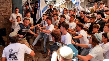 L'image montre un groupe animé de jeunes hommes dans une rue animée, probablement lors d'un événement ou d'une manifestation. Au premier plan, un homme portant un gilet "PRESS" semble être au centre de l'attention, entouré par plusieurs personnes qui cherchent à le repousser. Les participants portent des vêtements principalement blancs et bleu, suggérant une thématique nationale. L'atmosphère semble tendue, avec des expressions d'excitation et de conflit parmi la foule.
