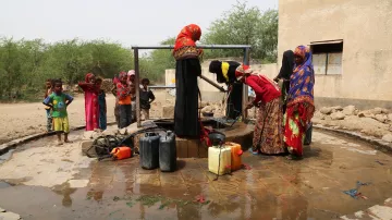 L'image montre un groupe de femmes près d'un puits. Elles sont vêtues de robes colorées et semblent être en train de remplir des conteneurs d'eau. Des enfants jouent autour d'elles. Le sol est humide, et le cadre semble rural, avec des buissons et une structure en arrière-plan. L'atmosphère semble active et communautaire, illustrant l'importance de l'accès à l'eau dans la vie quotidienne.