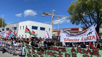L'image montre une manifestation avec une grande foule de personnes rassemblées. Les manifestants portent des bannières et des drapeaux, y compris des drapeaux palestiniens. Certains panneaux affichent des messages en faveur des droits du peuple palestinien et dénoncent le génocide à Gaza. L'ambiance semble être celle d'une protestation pacifique, avec des participants unis pour une cause commune. En arrière-plan, on peut voir des bâtiments et une grue de construction.