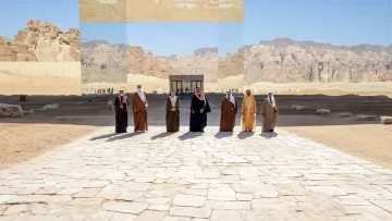 L'image montre un groupe d'individus en vêtements traditionnels, se tenant sur un chemin en pierre au milieu d'un paysage désertique. En arrière-plan, une grande structure réfléchissante fait un miroir du paysage environnant, créant un effet visuel intéressant. Les montagnes et le ciel sont également visibles, ajoutant une dimension naturelle à la scène. L'atmosphère semble solennelle et digne.