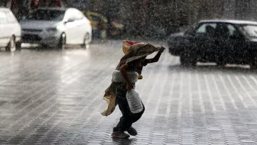 L'image montre un enfant courant sous la pluie intense, portant une écuelle en métal et un sac. Il semble se protéger de la pluie avec un tissu qui couvre sa tête et ses épaules. Le sol est mouillé et réfléchit la lumière, tandis que l'environnement autour de lui est flou en raison des cordes de pluie. Des voitures sont visibles en arrière-plan, accentuant l'atmosphère urbaine de la scène.