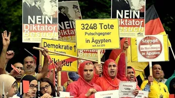 L'image montre un groupe de manifestants rassemblés pour une protestation. Ils portent des vêtements de pluie rouges et tiennent des pancartes avec des messages en allemand et en anglais, dénonçant une dictature et parlant de décès d'opposants en Égypte. On peut voir des slogans comme "NEIN" et des chiffres indiquant le nombre de morts. L'atmosphère semble sérieuse, témoignant de l'engagement des manifestants pour leurs causes.
