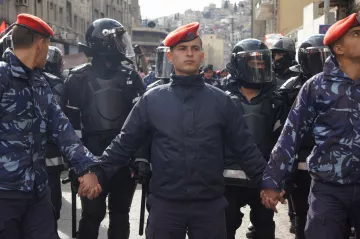L'image montre un groupe de policiers ou de soldats, avec des uniformes distinctifs, se tenant les mains. Au centre, un homme en majorette avec un béret se distingue. Derrière eux, des membres des forces de l'ordre portent des casques et des équipements de protection. L'environnement semble urbain, avec des bâtiments en arrière-plan. L'atmosphère peut suggérer une situation de tension ou de manifestation.