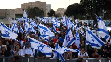 L'immagine mostra una folla di persone che partecipa a una manifestazione, sventolando molte bandiere di Israele. Il pubblico sembra essere molto numeroso e coinvolto, con alcuni partecipanti che usano telefoni cellulari. Sullo sfondo si possono vedere statue e edifici, mentre la scena è illuminata dalla luce del giorno. L'atmosfera appare vivace e carica di spirito collettivo.
