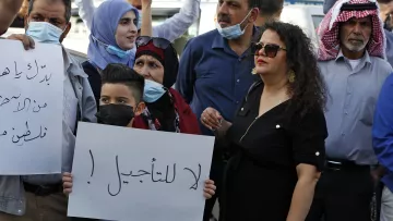 تظهر في الصورة مجموعة من الناس في مظاهرة. يرفع بعضهم لافتات، بينما يرتدي الآخرون كمامات. في المقدمة، يوجد طفل صغير يحمل لافتة مكتوب عليها "لا للتأجيل!"، وتحيط به مجموعة من البالغين، بينهم امرأة ترتدي فستانًا أسود وبعض الرجال بملابس تقليدية. تعكس الصورة جوًا من الاحتجاج والتعبير عن الرأي.