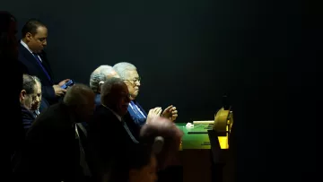 L'image montre un groupe d'hommes assis à une table, apparemment en train d'assister à un événement ou une réunion. L'éclairage est tamisé, mettant en évidence certaines personnes tandis que d'autres sont dans l'ombre. L'un des hommes est occupé avec son téléphone portable. Les visages semblent préoccupés et concentrés, donnant une impression de gravité et d'importance à la situation.