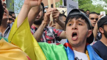 L'image montre une scène de manifestation, où un jeune homme, portant un drapeau coloré, crie avec passion. Il semble mobilisé et engagé dans la cause pour laquelle les manifestants se sont rassemblés. En arrière-plan, on peut voir d'autres personnes tenant des pancartes et exprimant leurs préoccupations. L'atmosphère est remplie d'énergie et de détermination.